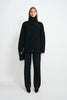 A model stands against a light gray studio background wearing a black high-neck button-front jacket with flap pockets, black wide-leg trousers, and black pointed shoes, while holding a small black handbag.