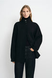 A model wearing a black high-neck button-front coat over a black top and dark jeans, standing against a plain light gray studio background.