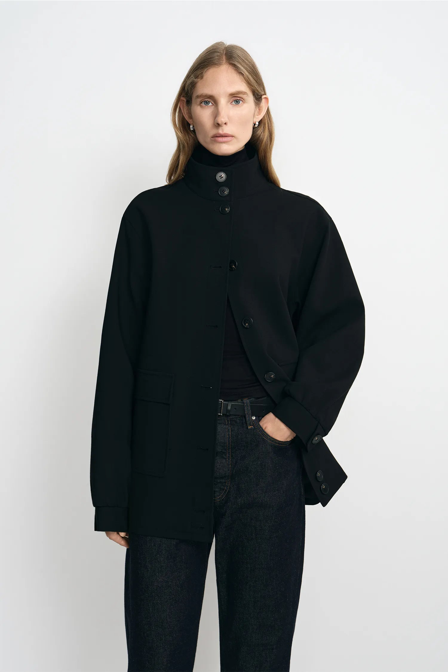 A model wearing a black high-neck button-front coat over a black top and dark jeans, standing against a plain light gray studio background.