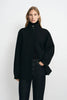 A model wearing a black high-neck button-front coat over a black top and dark jeans, standing against a plain light gray studio background.