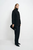 A model stands in profile wearing a black high-neck button-front coat, black wide-leg trousers, and black pointed shoes, while holding a black textured clutch against a plain light gray studio background.