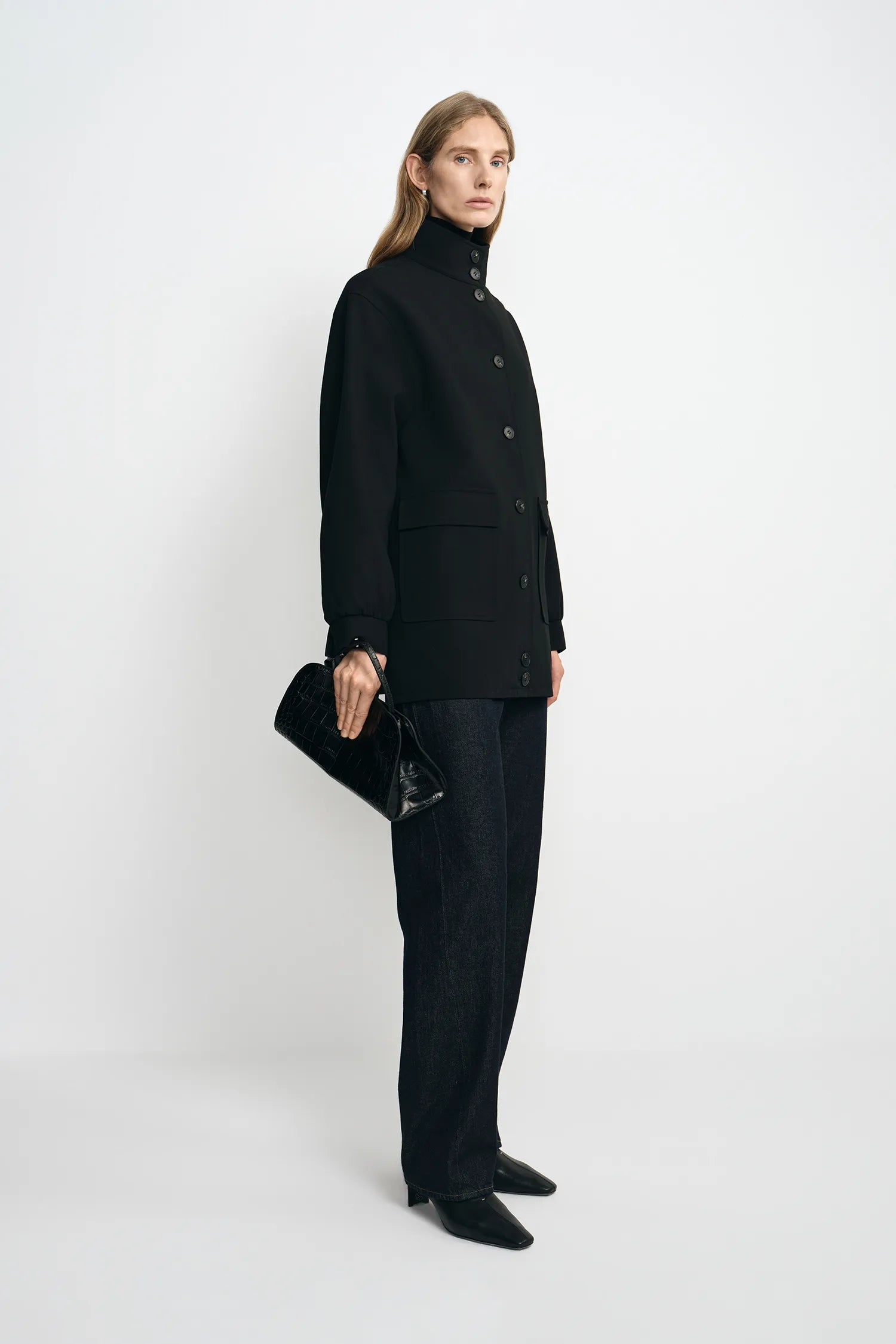 A model stands in profile wearing a black high-neck button-front coat, black wide-leg trousers, and black pointed shoes, while holding a black textured clutch against a plain light gray studio background.