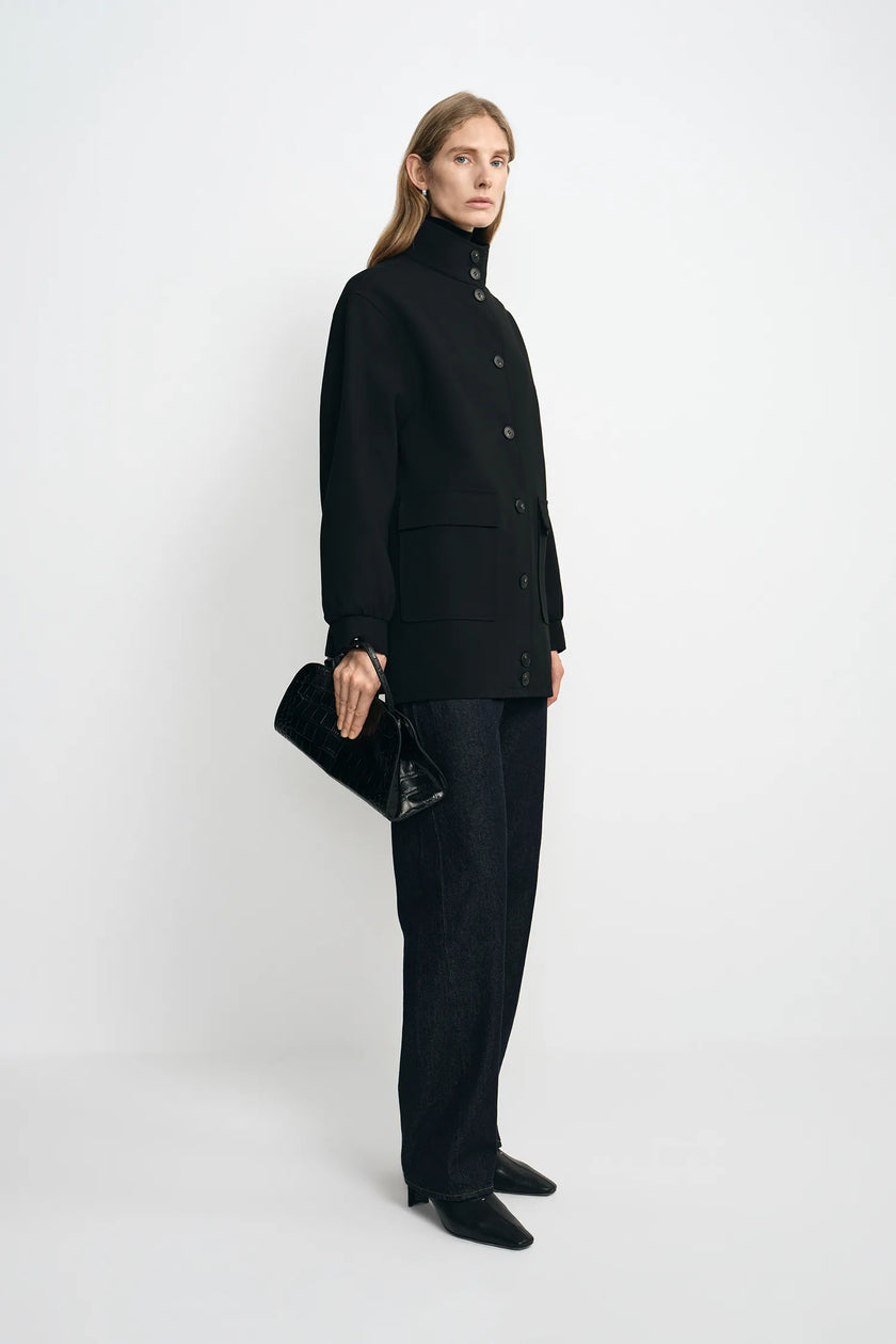 A model stands in profile wearing a black high-neck button-front coat, black wide-leg trousers, and black pointed shoes, while holding a black textured clutch against a plain light gray studio background.
