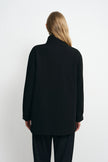 A woman viewed from the back wearing a black long-sleeve coat with a high collar and black pants, standing against a plain light gray studio background.