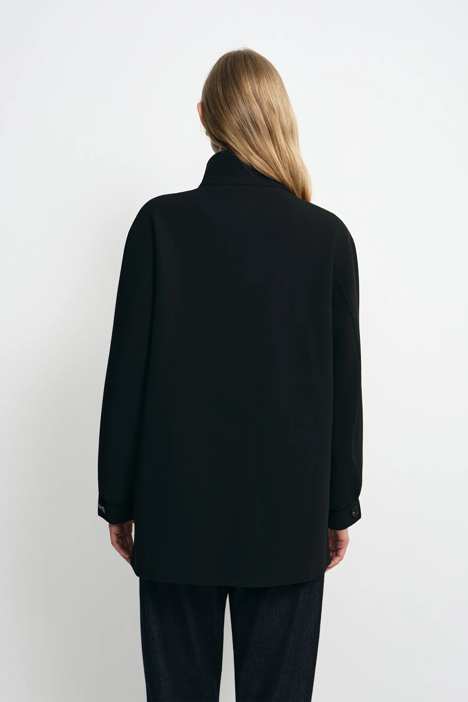 A woman viewed from the back wearing a black long-sleeve coat with a high collar and black pants, standing against a plain light gray studio background.