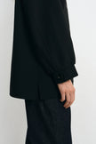 Close-up of a person wearing a black long-sleeve blazer with a buttoned cuff and dark textured trousers, shown against a plain light background.