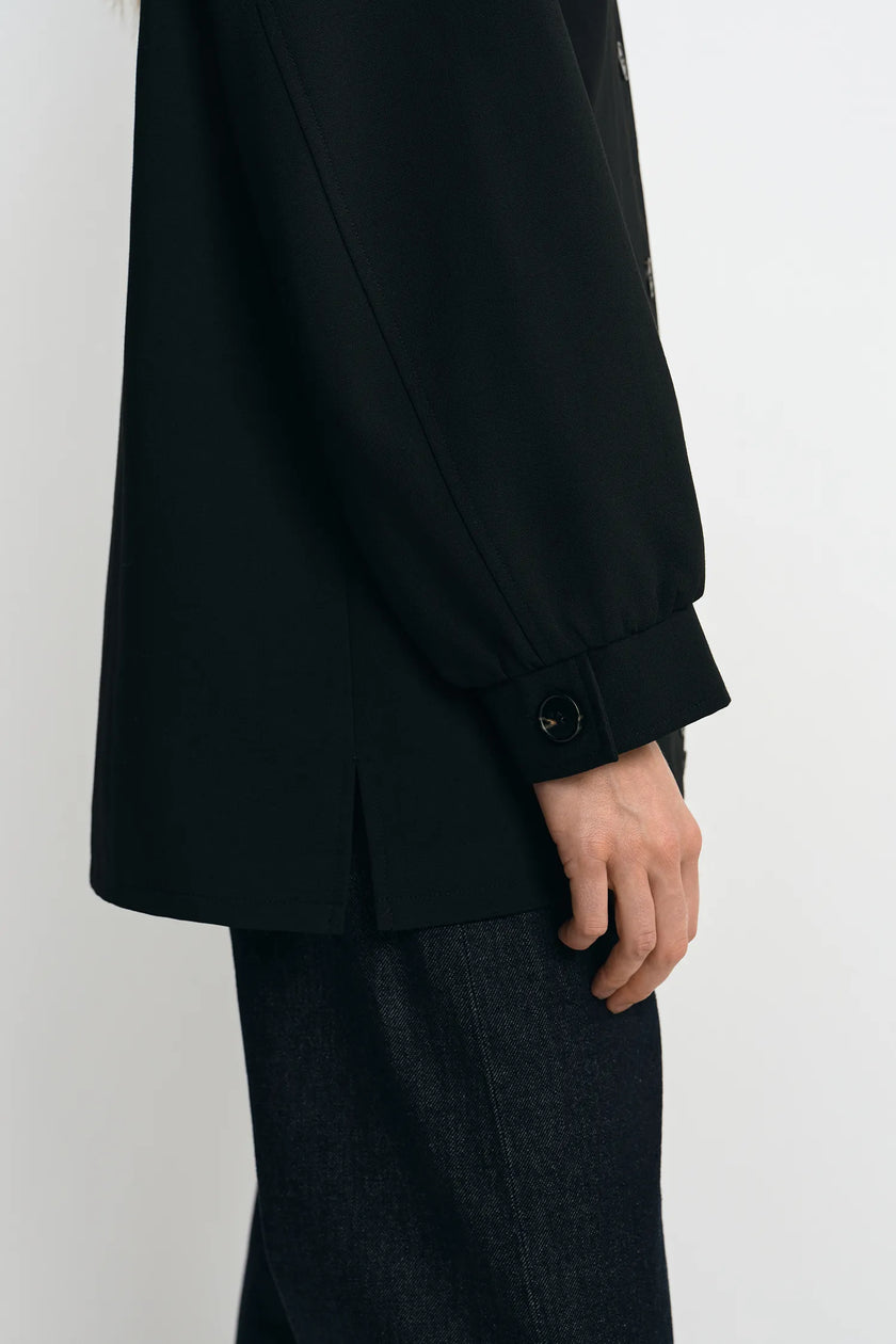 Close-up of a person wearing a black long-sleeve blazer with a buttoned cuff and dark textured trousers, shown against a plain light background.
