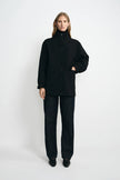 A model stands against a light gray studio background wearing a black high-neck button-front jacket with long sleeves, black wide-leg pants, and black shoes.