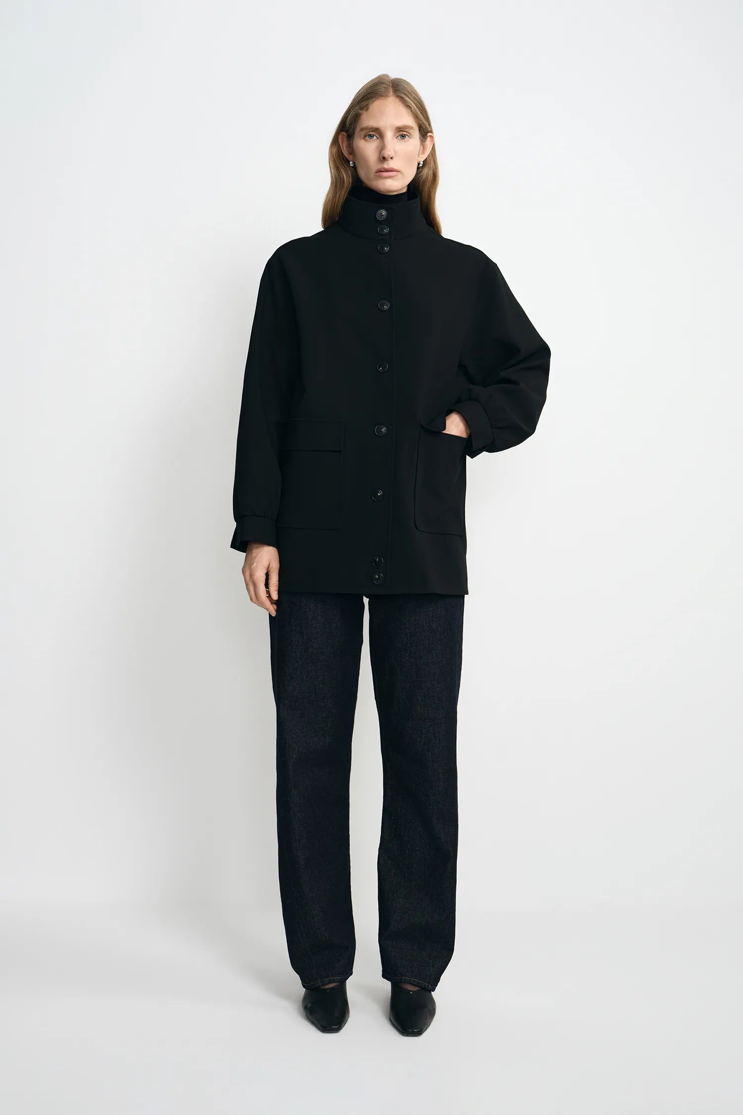 A model stands against a light gray studio background wearing a black high-neck button-front jacket with long sleeves, black wide-leg pants, and black shoes.