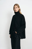 A model wearing a black high-neck button-front coat with wide sleeves, paired with dark jeans, photographed against a plain light gray studio background.