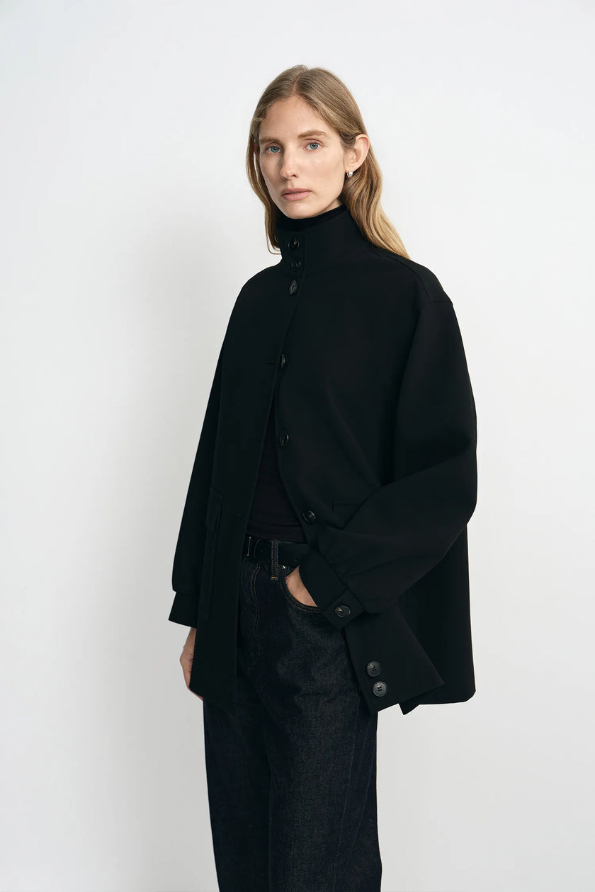A model wearing a black high-neck button-front coat with wide sleeves, paired with dark jeans, photographed against a plain light gray studio background.