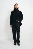 A model stands against a plain white studio background wearing a black high-neck button-front coat over a black top and dark wide-leg jeans.