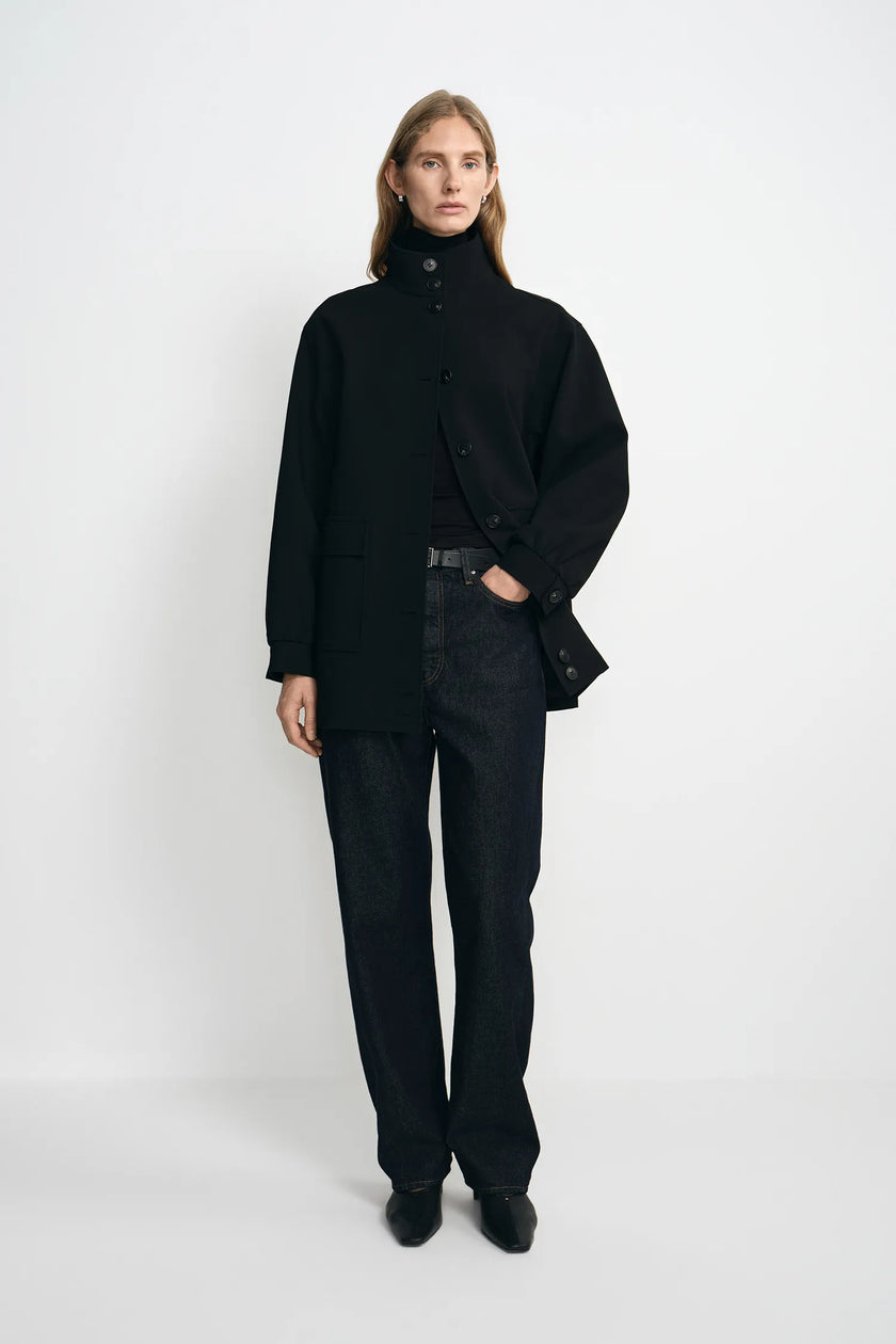A model stands against a plain white studio background wearing a black high-neck button-front coat over a black top and dark wide-leg jeans.