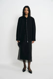 A model stands against a plain light gray studio background wearing a black long-sleeve jacket with a front zipper over a black midi skirt with sheer overlay, black tights, and black pointed shoes.