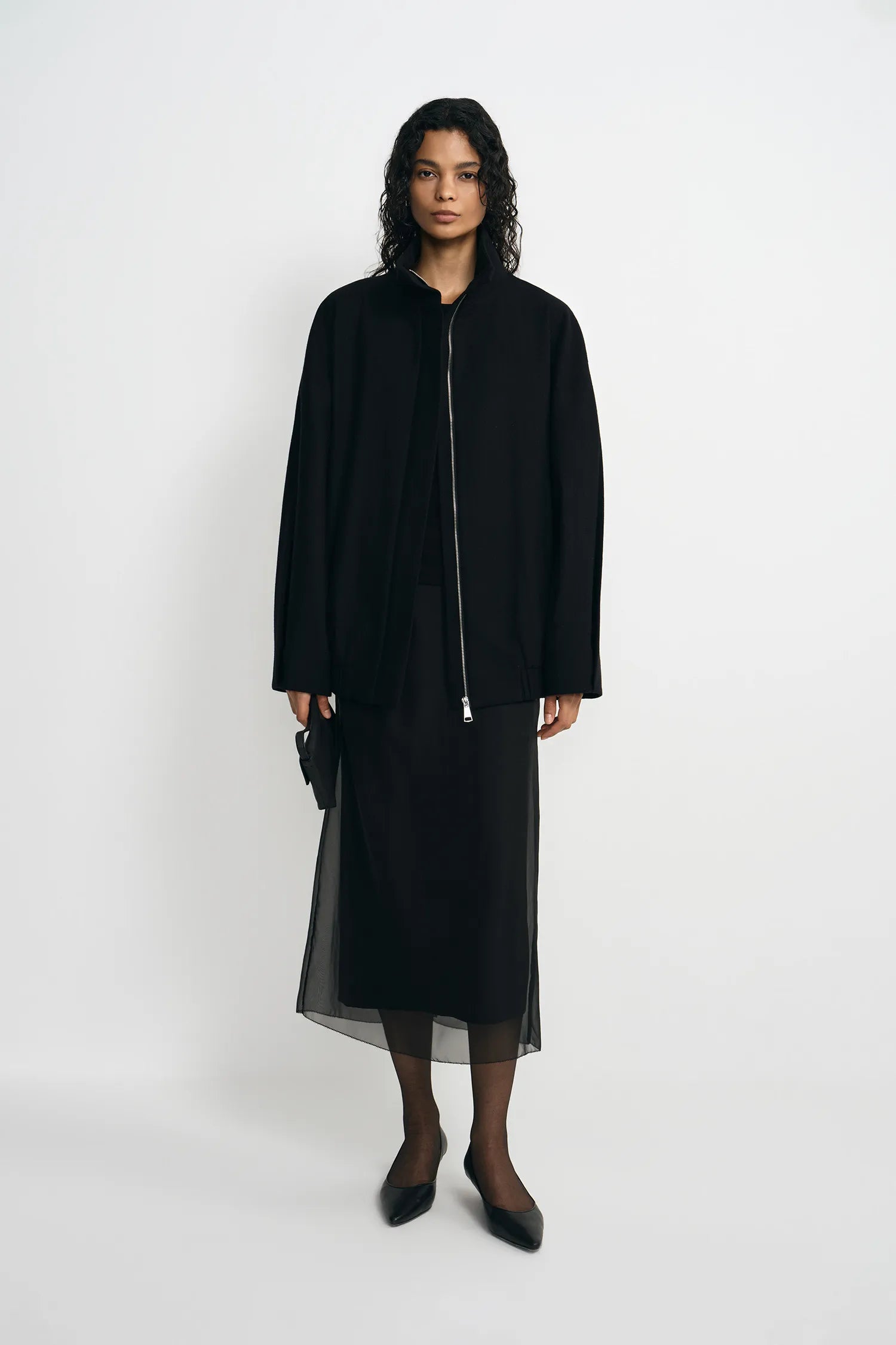 A model stands against a plain light gray studio background wearing a black long-sleeve jacket with a front zipper over a black midi skirt with sheer overlay, black tights, and black pointed shoes.
