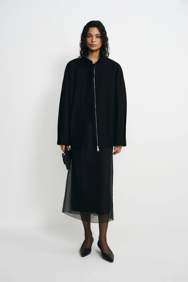 A model stands against a plain light gray studio background wearing a black long-sleeve jacket with a front zipper over a black midi skirt with sheer overlay, black tights, and black pointed shoes.