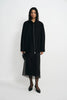 A model stands against a plain light gray studio background wearing a black long-sleeve jacket with a front zipper over a black midi skirt with sheer overlay, black tights, and black pointed shoes.