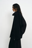 A model with long, curly dark hair is shown in profile wearing a black oversized zip-up jacket and a matching black skirt against a plain white studio background.