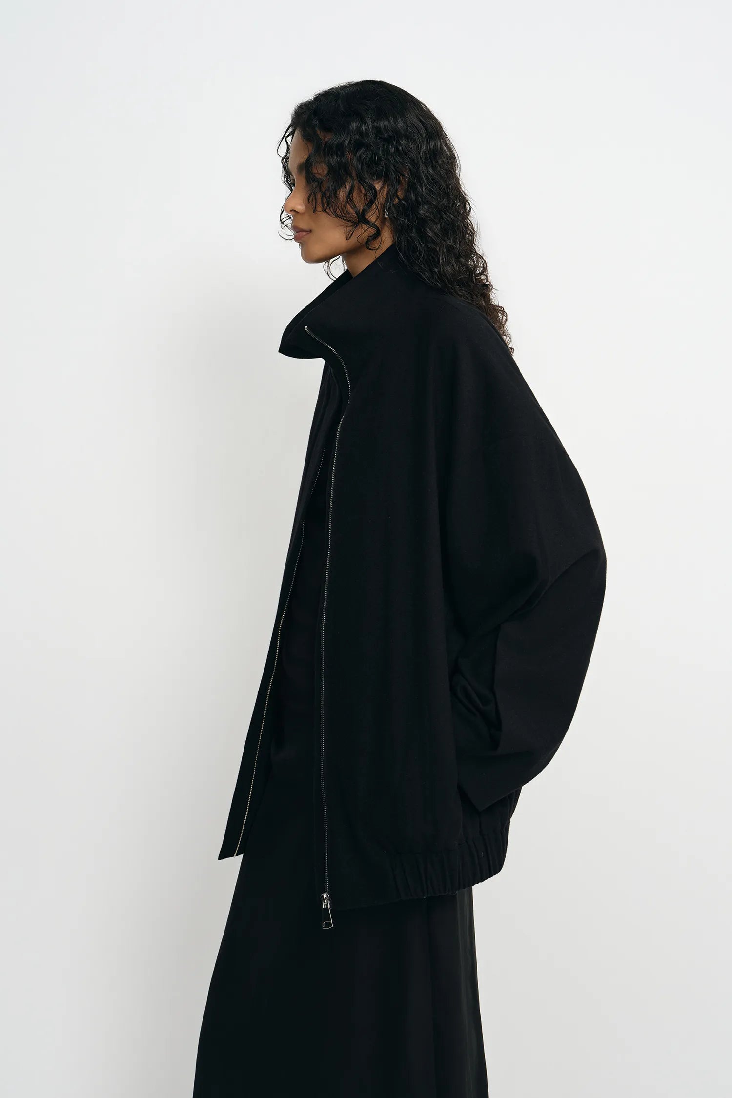 A model with long, curly dark hair is shown in profile wearing a black oversized zip-up jacket and a matching black skirt against a plain white studio background.