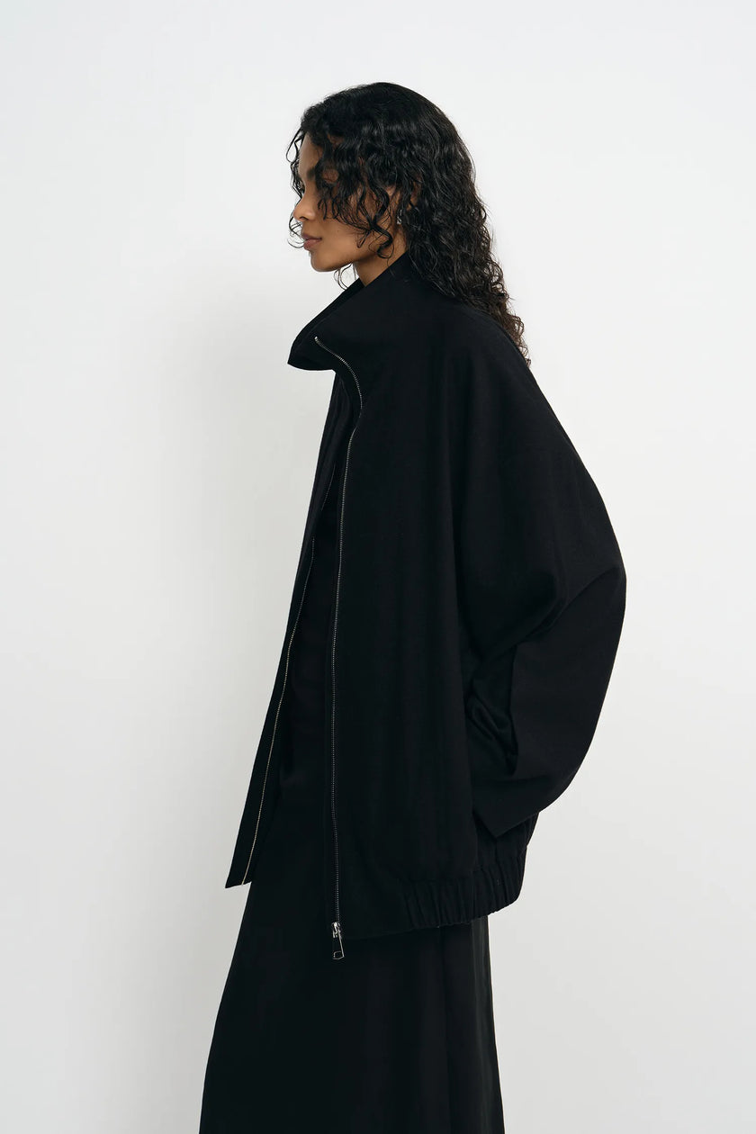 A model with long, curly dark hair is shown in profile wearing a black oversized zip-up jacket and a matching black skirt against a plain white studio background.