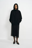 A model stands against a plain white studio background wearing a black high-neck long-sleeve top with a sheer black midi skirt and black pointed shoes.