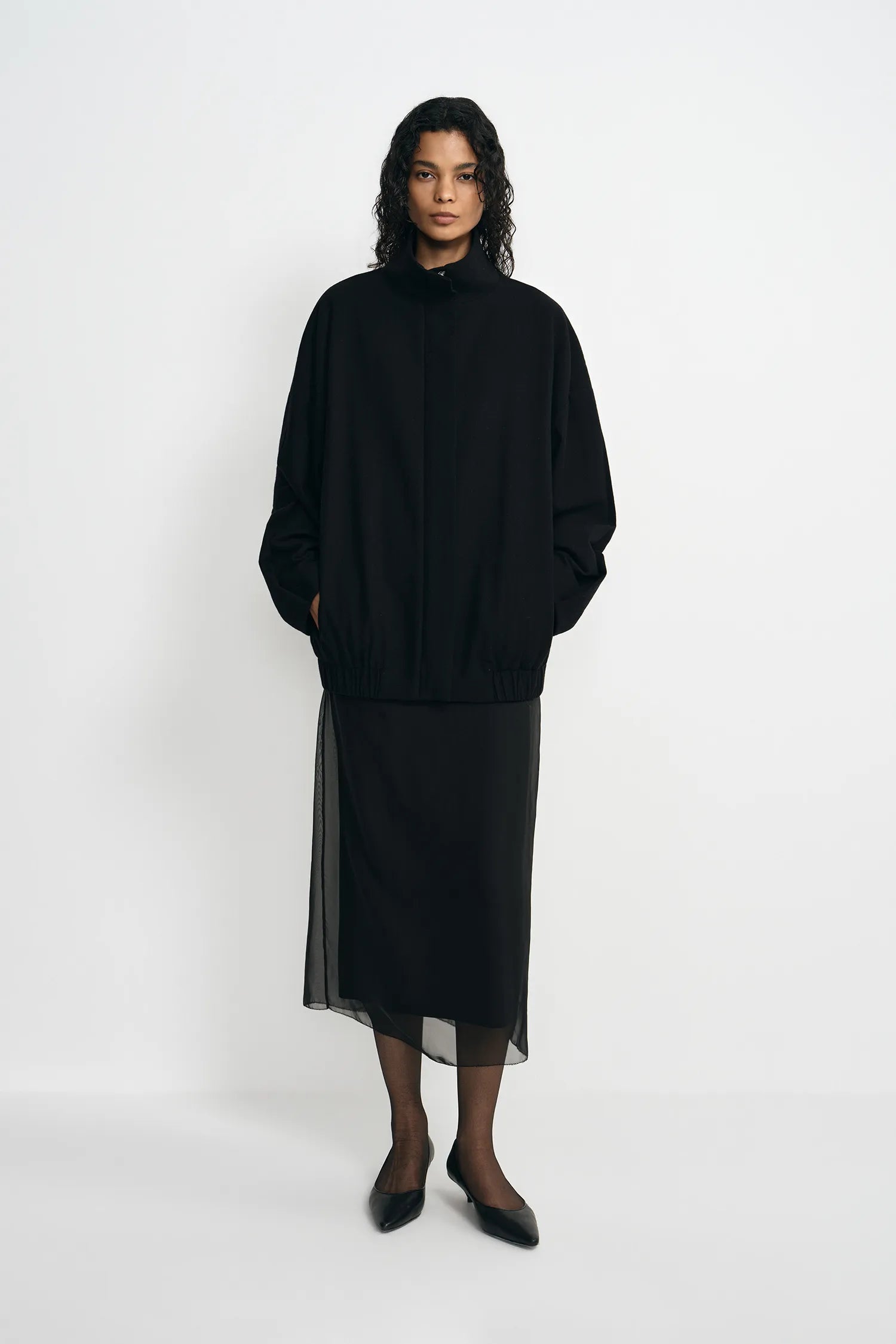 A model stands against a plain white studio background wearing a black high-neck long-sleeve top with a sheer black midi skirt and black pointed shoes.