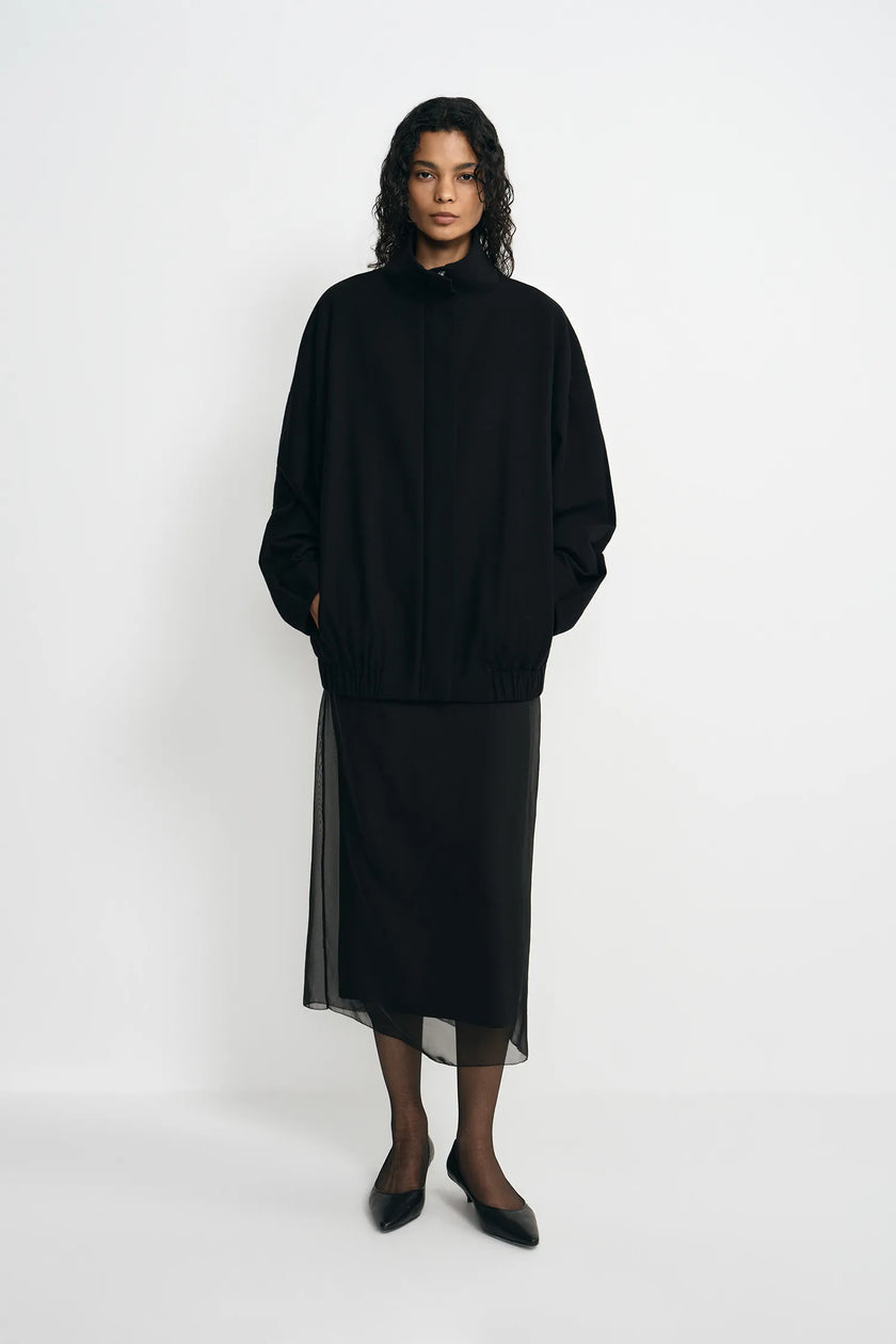 A model stands against a plain white studio background wearing a black high-neck long-sleeve top with a sheer black midi skirt and black pointed shoes.