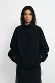 A model with curly dark hair wearing a black oversized mock-neck jacket with a drawstring and a black skirt, standing against a light gray studio background.