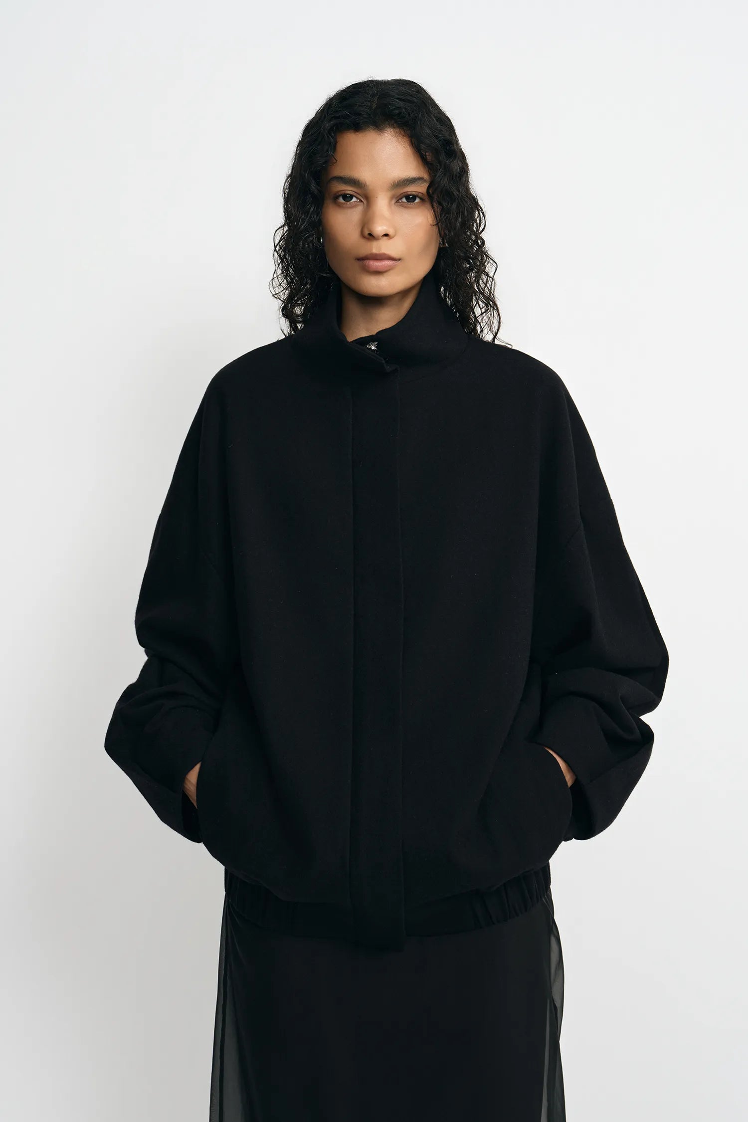 A model with curly dark hair wearing a black oversized mock-neck jacket with a drawstring and a black skirt, standing against a light gray studio background.