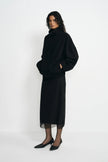 A model wearing a black turtleneck sweater and a matching black midi skirt with sheer black tights and black pointed shoes, standing against a plain white studio background.
