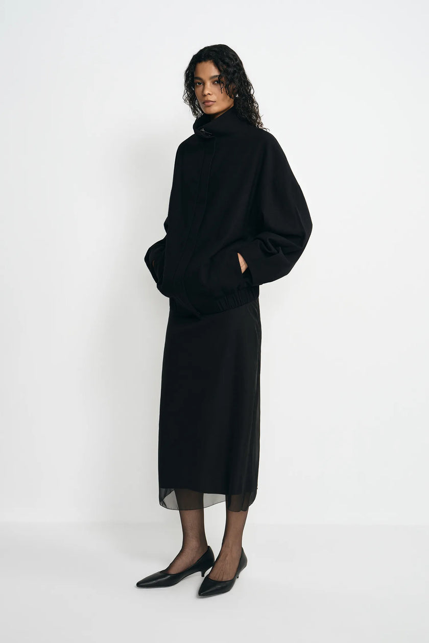 A model wearing a black turtleneck sweater and a matching black midi skirt with sheer black tights and black pointed shoes, standing against a plain white studio background.