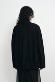 A person shown from the back wearing a loose black knit sweater and a sheer black skirt against a plain white studio background.