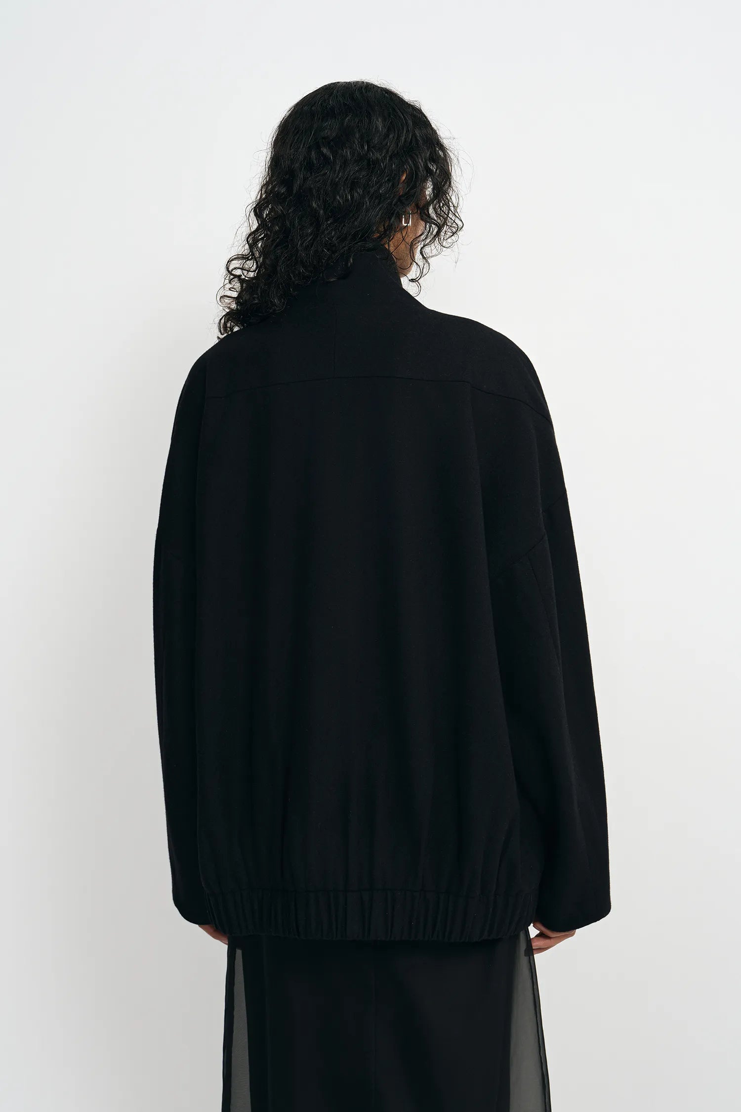 A person shown from the back wearing a loose black knit sweater and a sheer black skirt against a plain white studio background.