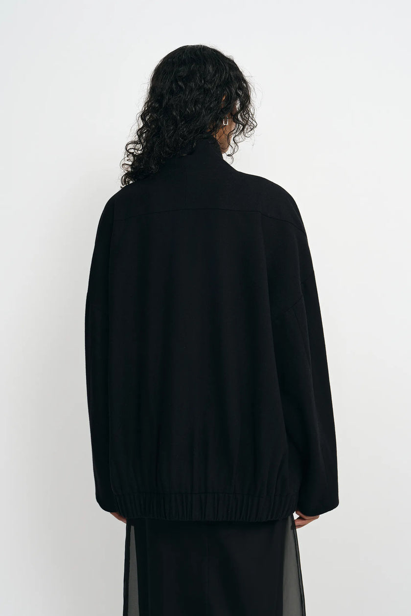 A person shown from the back wearing a loose black knit sweater and a sheer black skirt against a plain white studio background.