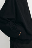 Close-up of a person wearing a black oversized long-sleeve top or jacket with a gathered elastic waist, shown against a plain light background.