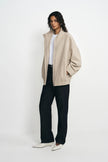 A model stands against a plain white studio background wearing a light beige oversized zip-up cardigan over a white top, paired with dark charcoal wide-leg pants and white pointed flats.