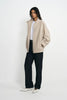 A model stands against a plain white studio background wearing a light beige oversized zip-up cardigan over a white top, paired with dark charcoal wide-leg pants and white pointed flats.