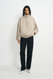 A model stands against a plain white studio background wearing a light beige textured jacket with a high mock neck, dark charcoal pants, and white pointed shoes.