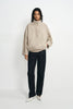 A model stands against a plain white studio background wearing a light beige textured jacket with a high mock neck, dark charcoal pants, and white pointed shoes.