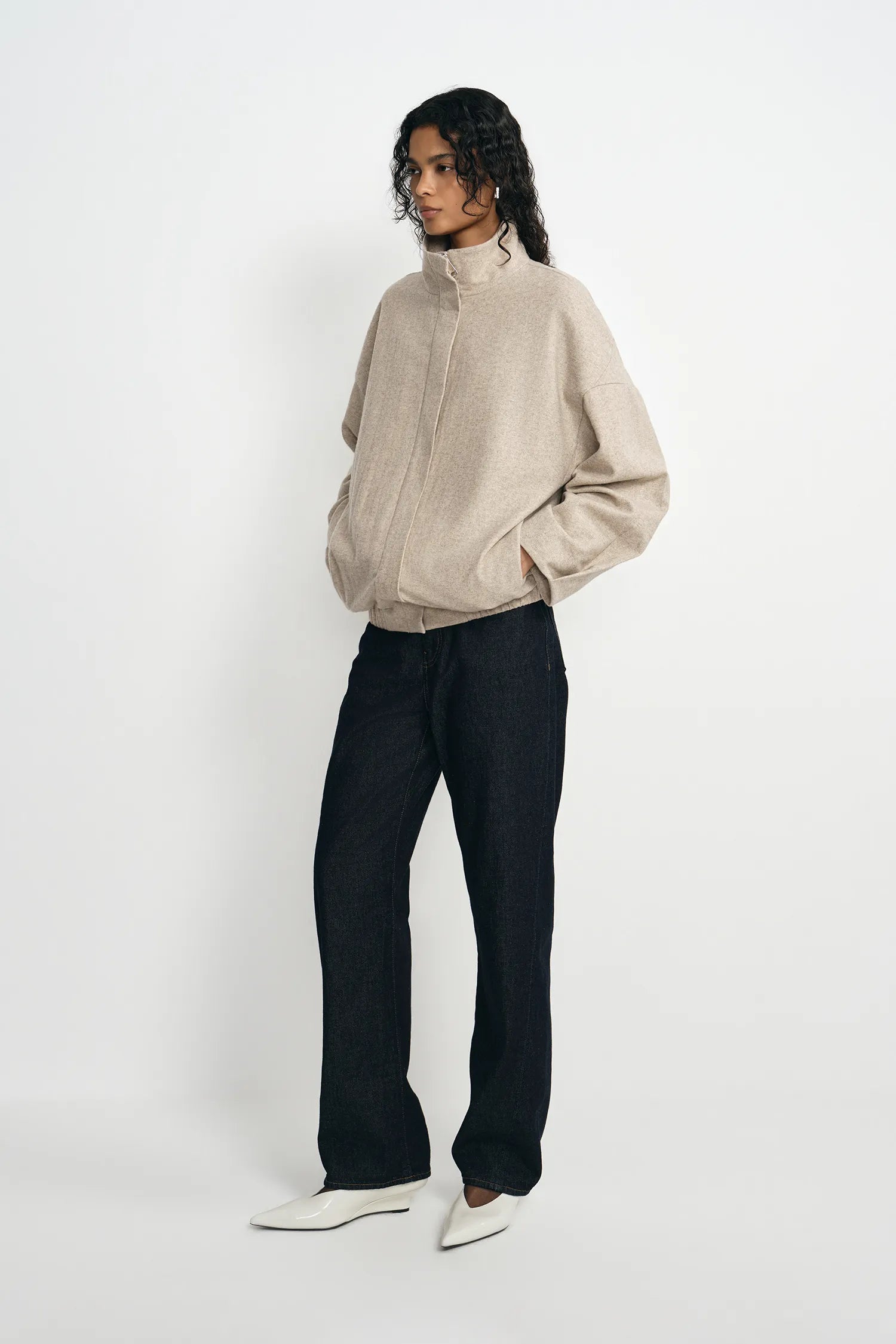 A model stands against a plain white studio background wearing a light beige textured zip-up sweater, dark charcoal wide-leg jeans, and white pointed-toe flats.