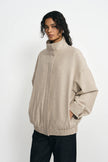 A model wearing a light beige knit zip-up jacket with a mock neck and oversized fit, paired with dark pants, against a plain white studio background.