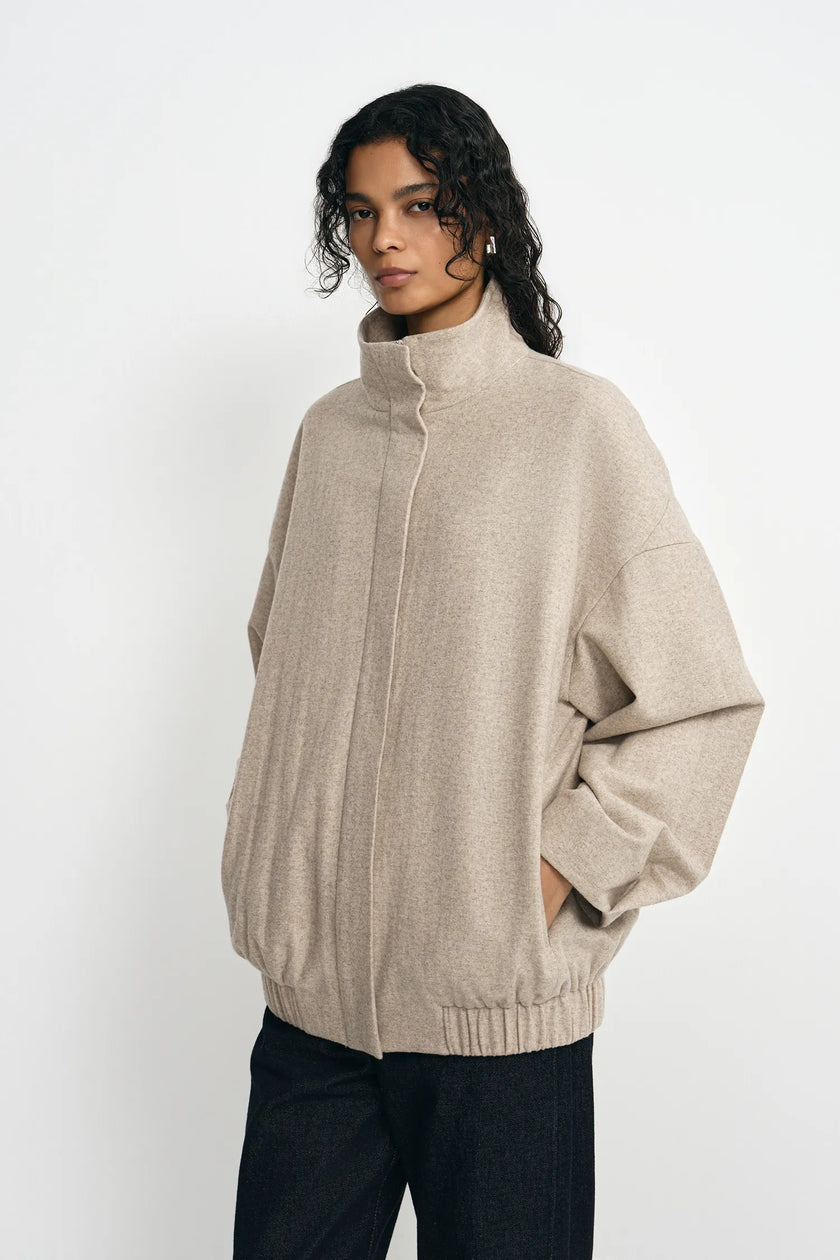 A model wearing a light beige knit zip-up jacket with a mock neck and oversized fit, paired with dark pants, against a plain white studio background.