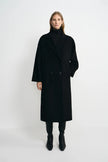 A model stands against a light gray studio background wearing a long black double-breasted coat over a black turtleneck, with black pants and black ankle boots.