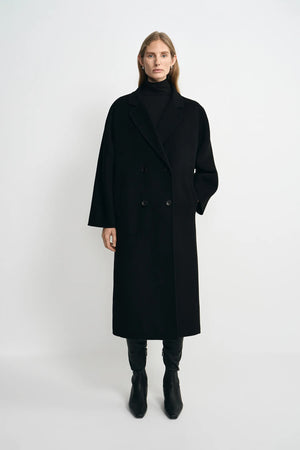 A model stands against a light gray studio background wearing a long black double-breasted coat over a black turtleneck, with black pants and black ankle boots.
