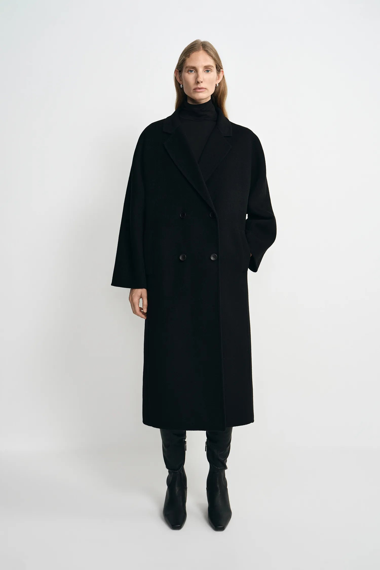 A model stands against a light gray studio background wearing a long black double-breasted coat over a black turtleneck, with black pants and black ankle boots.
