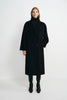 A model stands against a light gray studio background wearing a long black double-breasted coat over a black turtleneck, with black pants and black ankle boots.