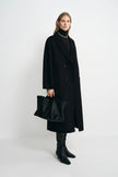 A model stands in a studio wearing a long black double-breasted coat over a black turtleneck, with black knee-high boots and a black leather slouchy handbag.
