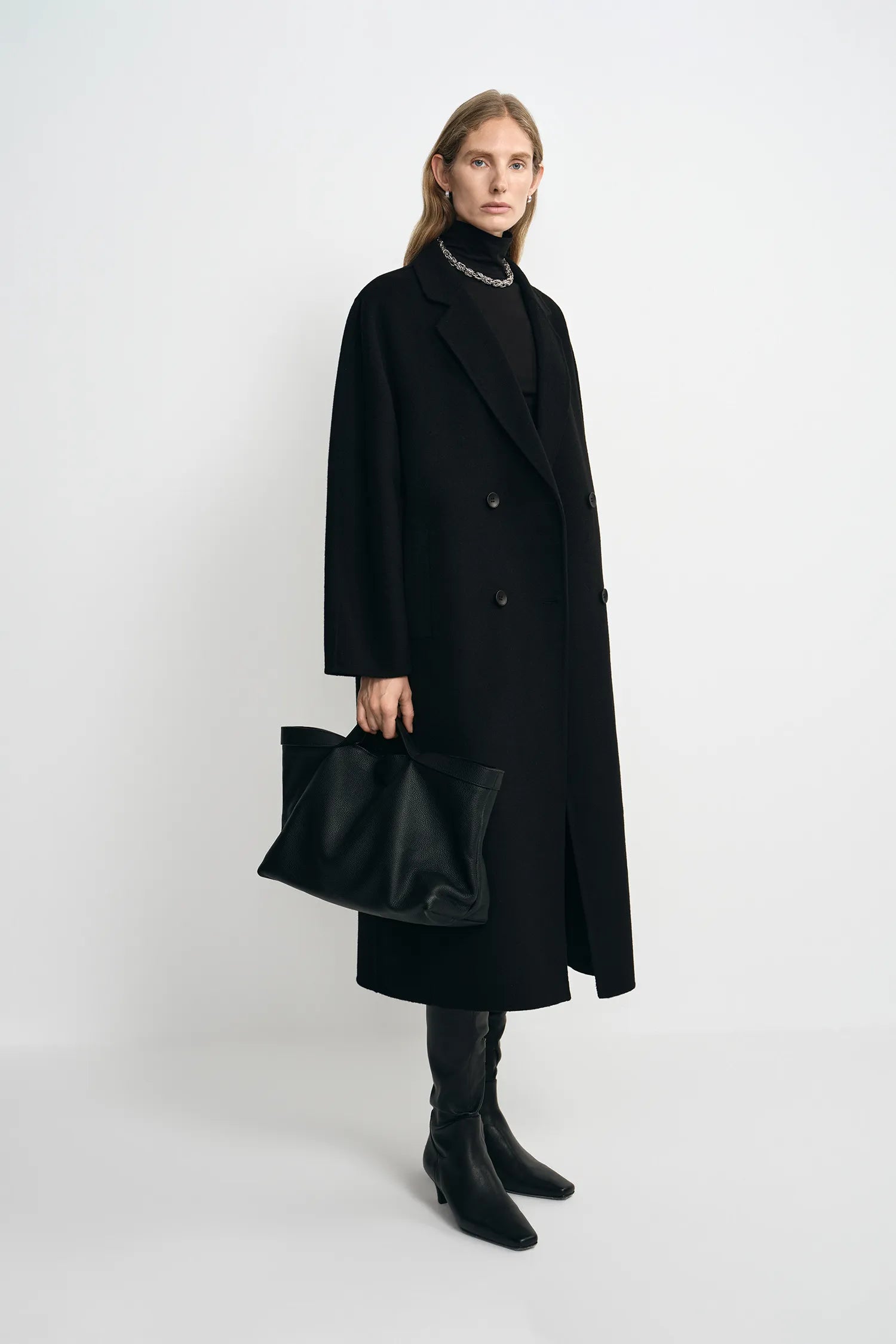 A model stands in a studio wearing a long black double-breasted coat over a black turtleneck, with black knee-high boots and a black leather slouchy handbag.