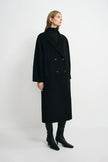 A woman modeling a long black double-breasted coat over a black turtleneck sweater, paired with black boots, against a plain light gray studio background.