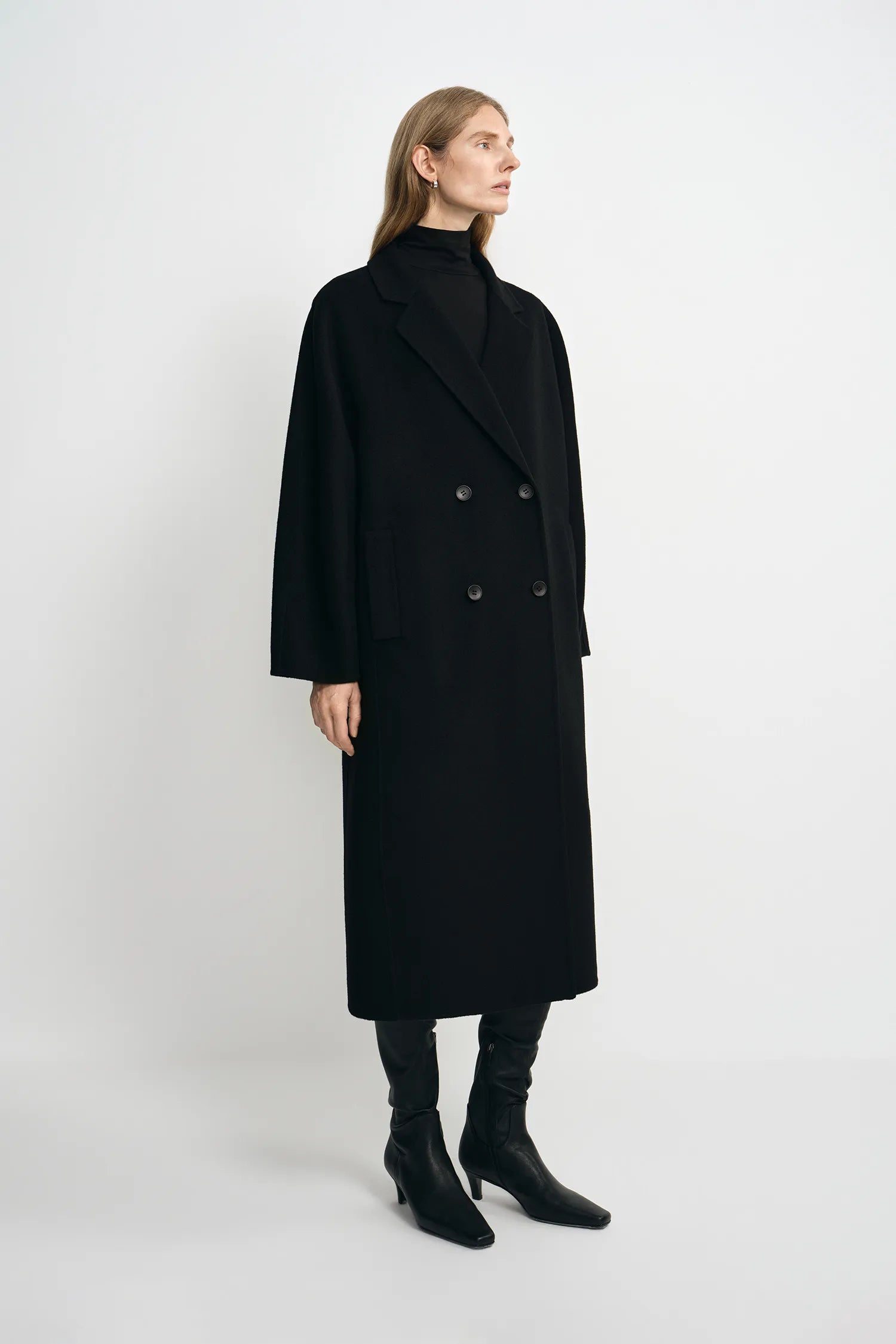 A woman modeling a long black double-breasted coat over a black turtleneck sweater, paired with black boots, against a plain light gray studio background.